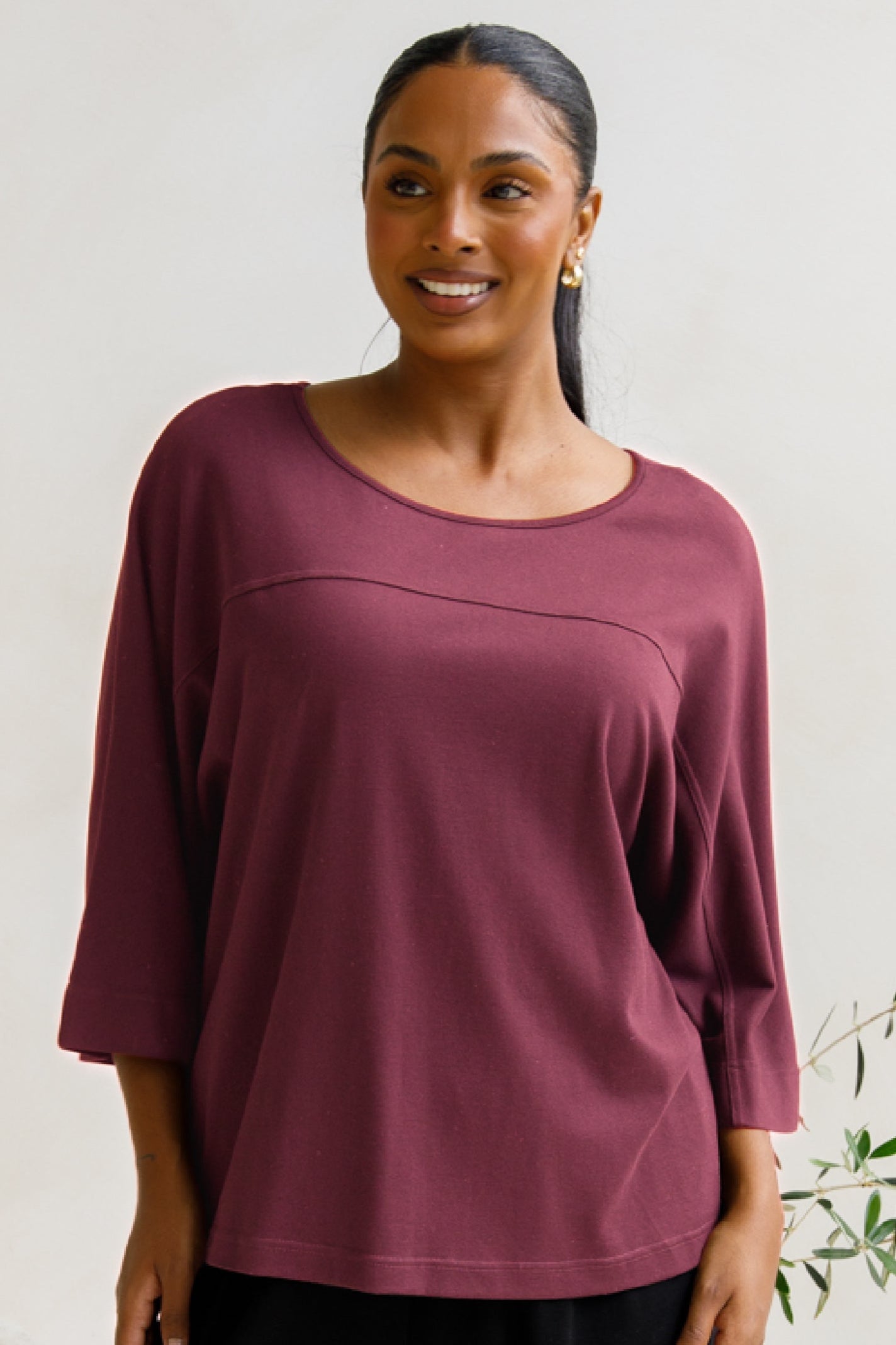 A woman standing in a lounge setting wearing a maroon, relaxed fit lounge top with 3/4 length sleeves and black lounge pants.