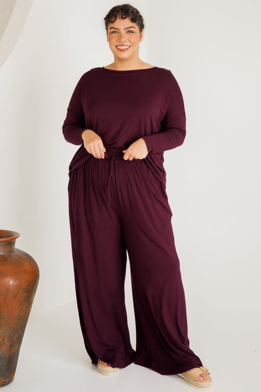 Woman wearing a burgundy jumpsuit standing next to a vase on a white background